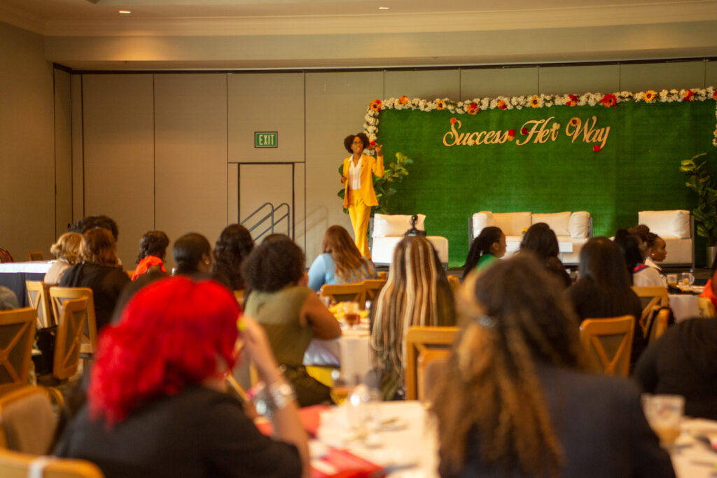 Emerald Sparks speaking onstage in yellow pants suit to women in audience